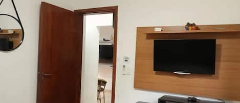Une pièce avec une porte en bois, un téléviseur à écran plat fixé au mur, une petite étagère au-dessus du téléviseur et une chaise au fond de la pièce.