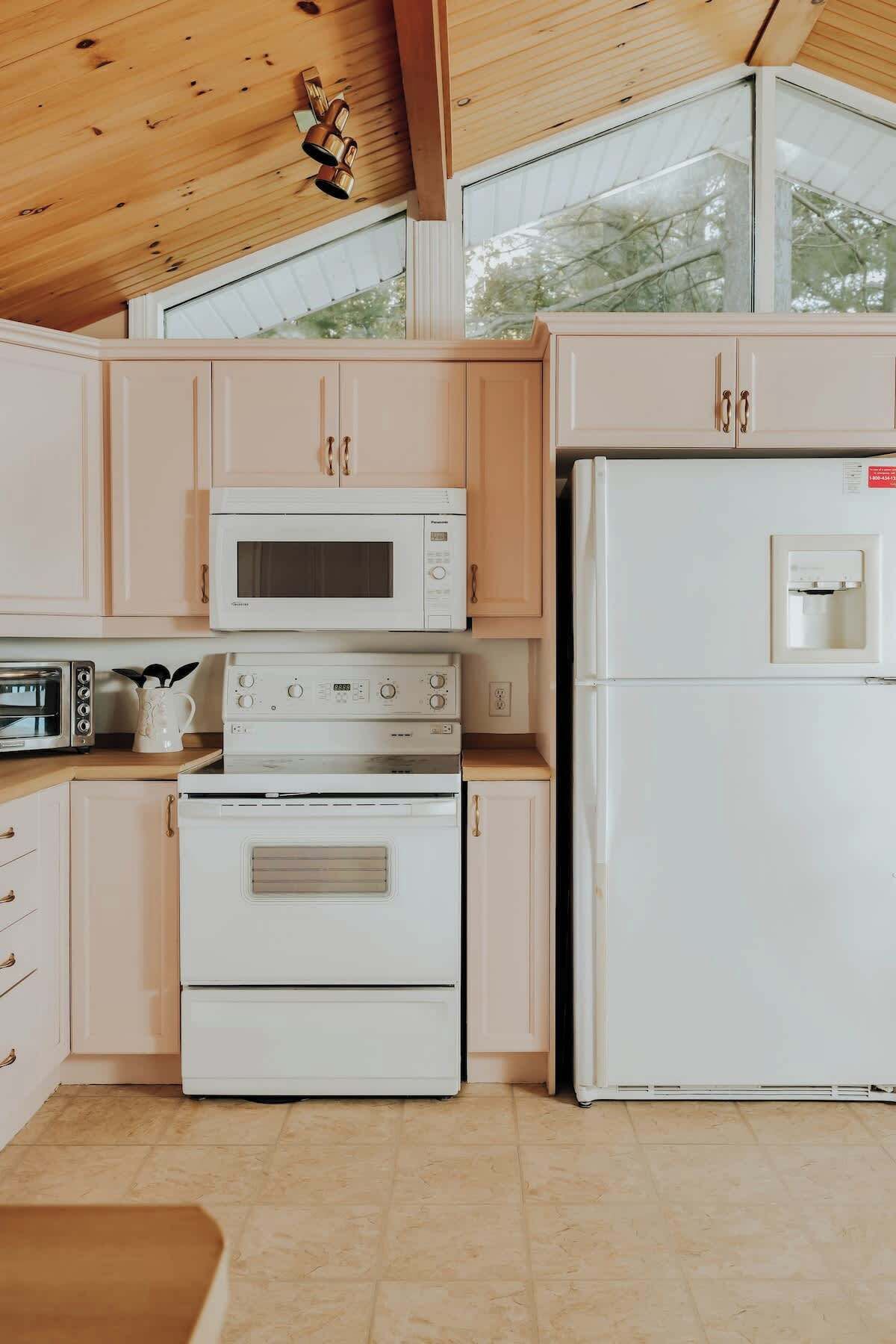 A kitchen with a microwave, oven, and refrigerator, wooden cabinets, and a window with a view of trees.