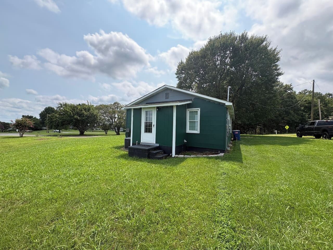 A small green house with a white door and windows, surrounded by a grassy field and trees.