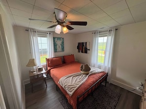 A bedroom with a bed, a ceiling fan, a nightstand with a lamp, and a window with curtains.