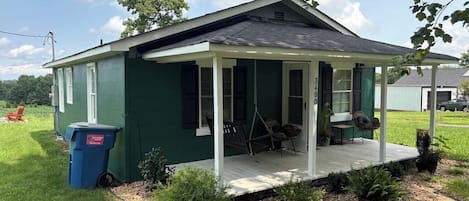 Une petite maison à un seul étage, dotée d’un porche, d’une balançoire et d’une poubelle bleue.