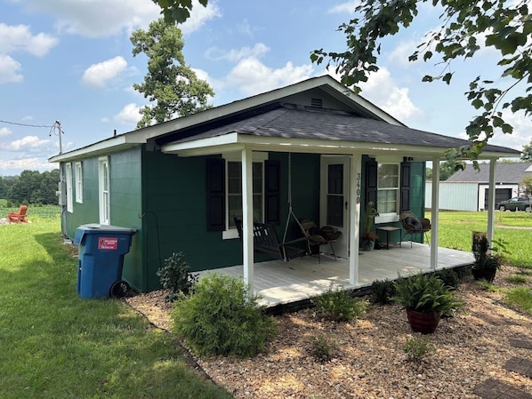 A small, single-story house with a porch, a swing chair, and a blue trash bin.