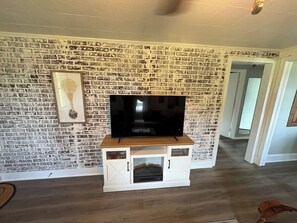 A living room with a brick-patterned wall, a flat-screen TV on a wooden stand, and a white entertainment unit with storage. - Cozy Country Cottage | 10 Min to Downtown Elkin (Elkin)