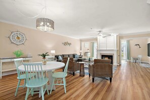 A spacious living room with a fireplace, a dining area with a round table and chairs, and a kitchen area with white cabinets. - Large Waterfront Townhome with Great Amenities (Ocean City)