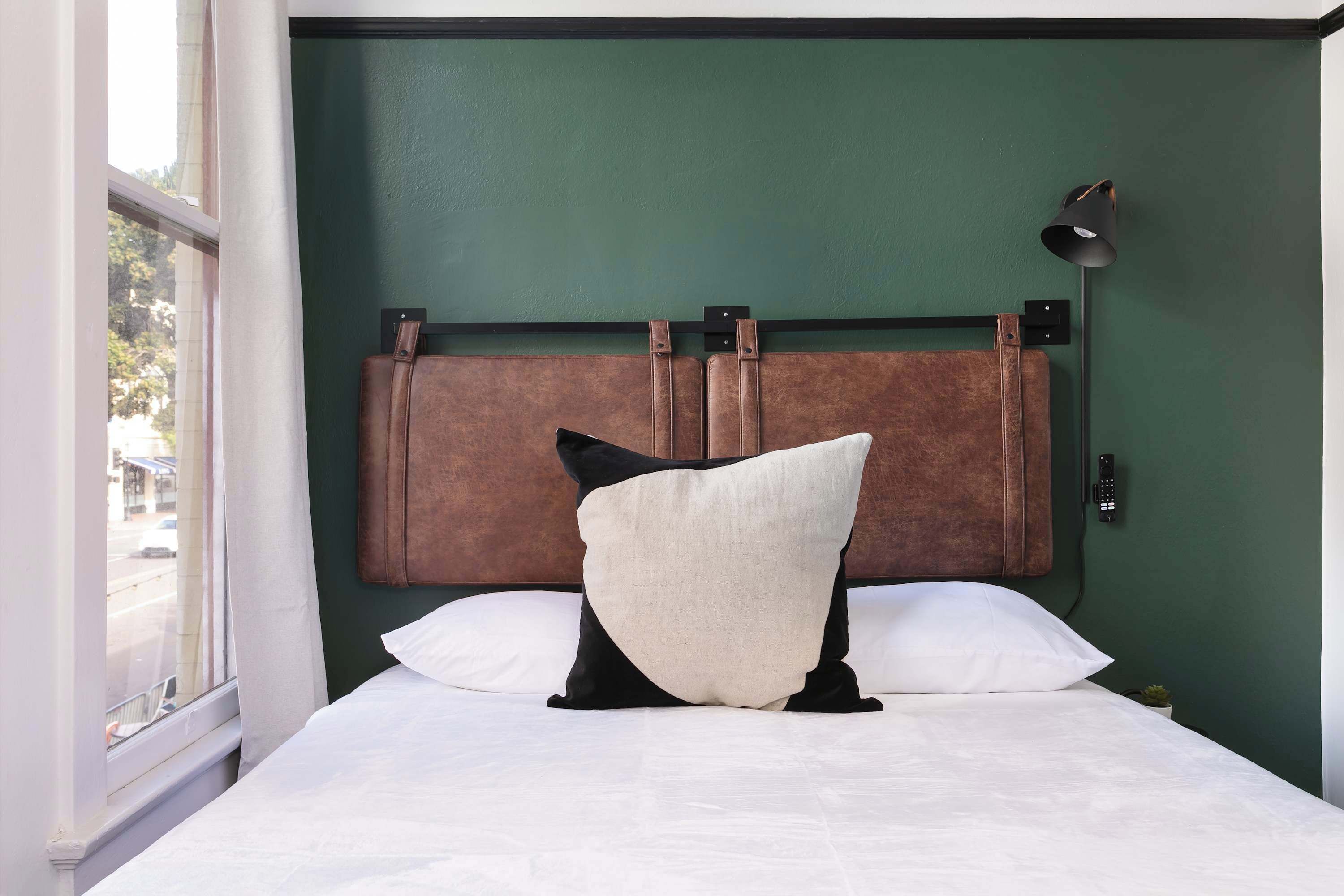 A bed with a headboard against a green wall, a window on the left, and a wall-mounted lamp.