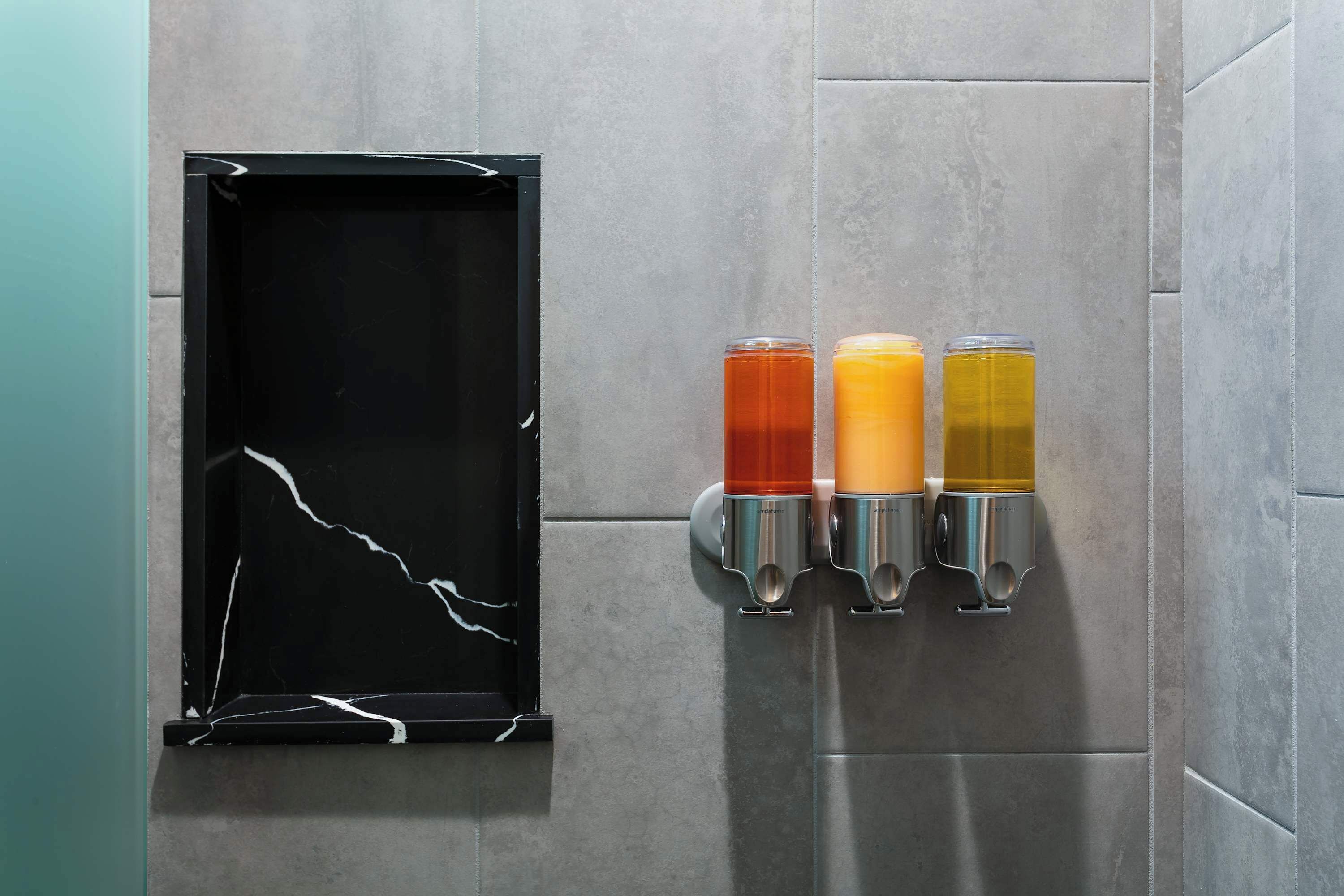 A wall-mounted beverage dispenser with three different colored drinks, a black-framed mirror, and a tiled wall.