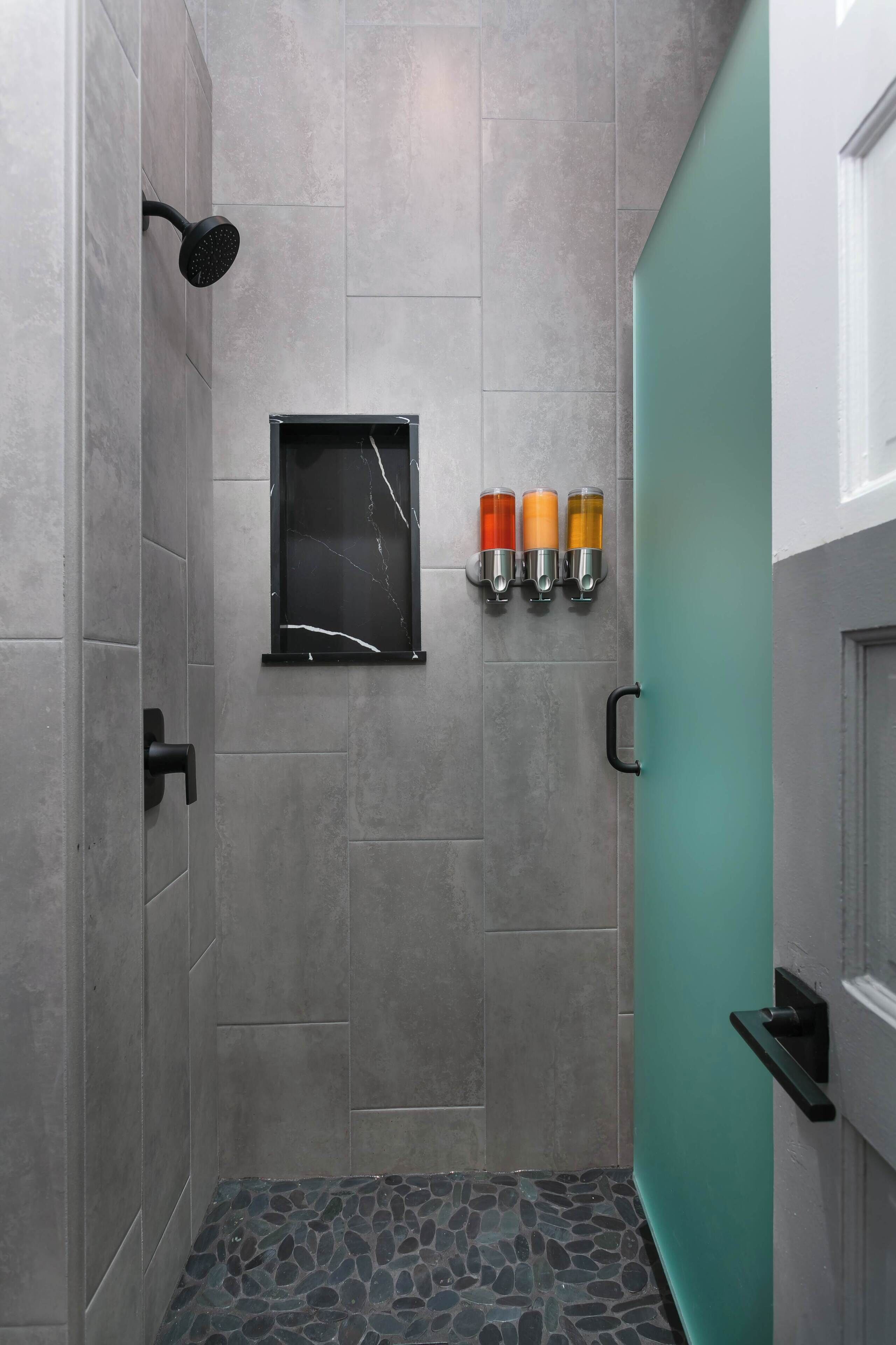 Une douche avec une porte en verre, une étagère noire fixée au mur avec trois bouteilles, et un sol carrelé.