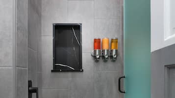 Um chuveiro com porta de vidro, uma prateleira preta fixada na parede com três frascos, e um chão de azulejos.