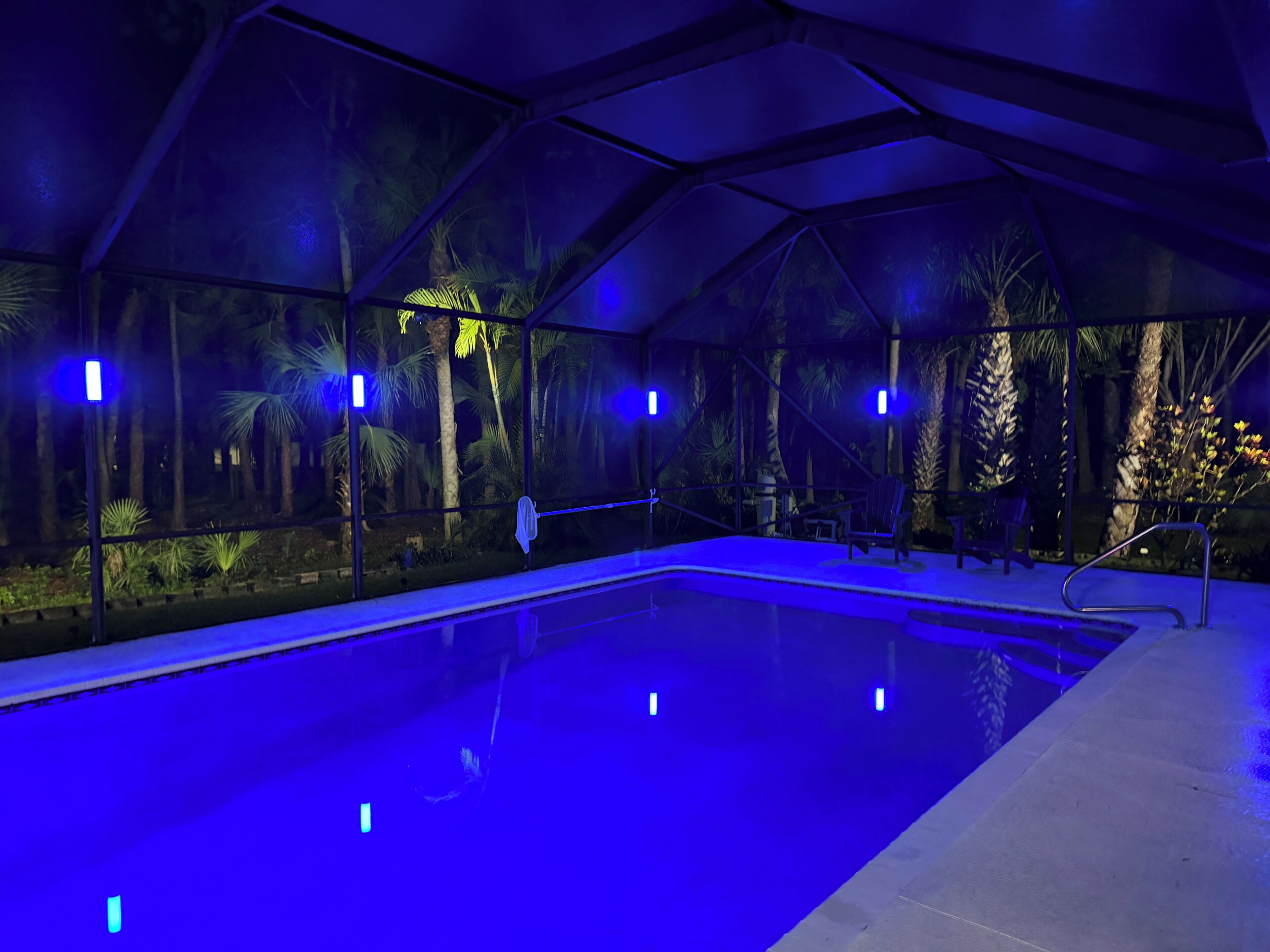 An indoor swimming pool with clear water, surrounded by glass walls and palm trees, under blue lighting.