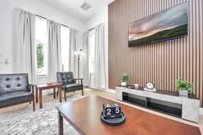 A modern living room with a sofa, a chair, a coffee table, a TV mounted on a wooden wall, and a calendar on the table. - Château Idaho - Cozy•Sleeps 10•NRG•Med Center•UoH (Houston)