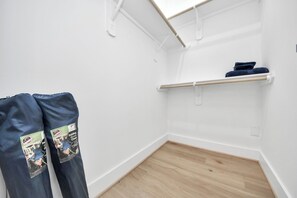A room with a white wall, a wooden floor, a white shelf, and a pair of dark blue pants hanging on a hook. - Château Idaho - Cozy•Sleeps 10•NRG•Med Center•UoH (Houston)