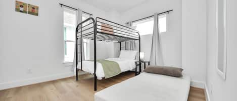 A room with a bunk bed, a single bed, a ceiling fan, and a window with curtains.