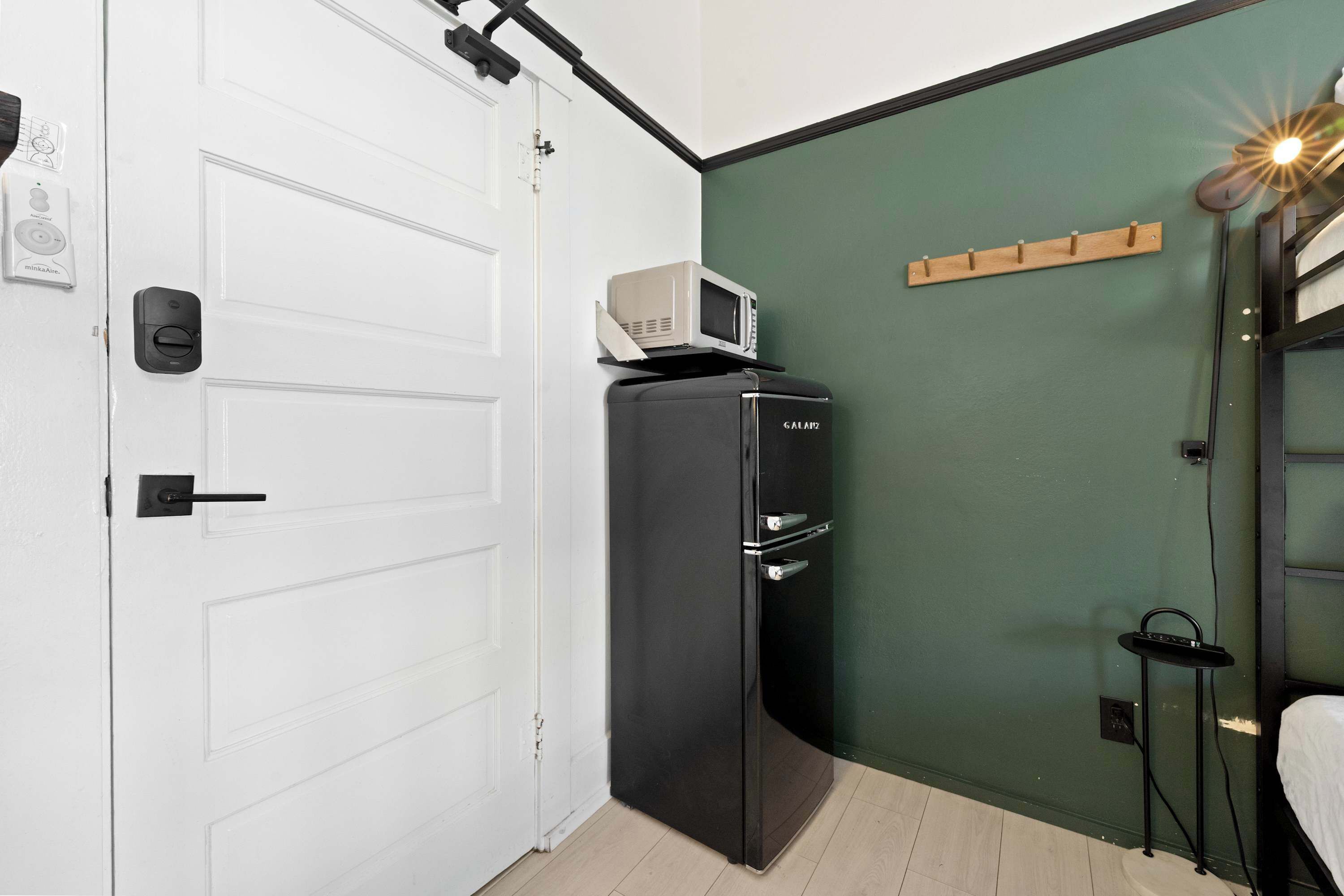 Une pièce de taille modeste avec une porte blanche, un réfrigérateur noir, un four à micro-ondes posé sur le dessus, un mur vert et un lit avec une tête de lit.
