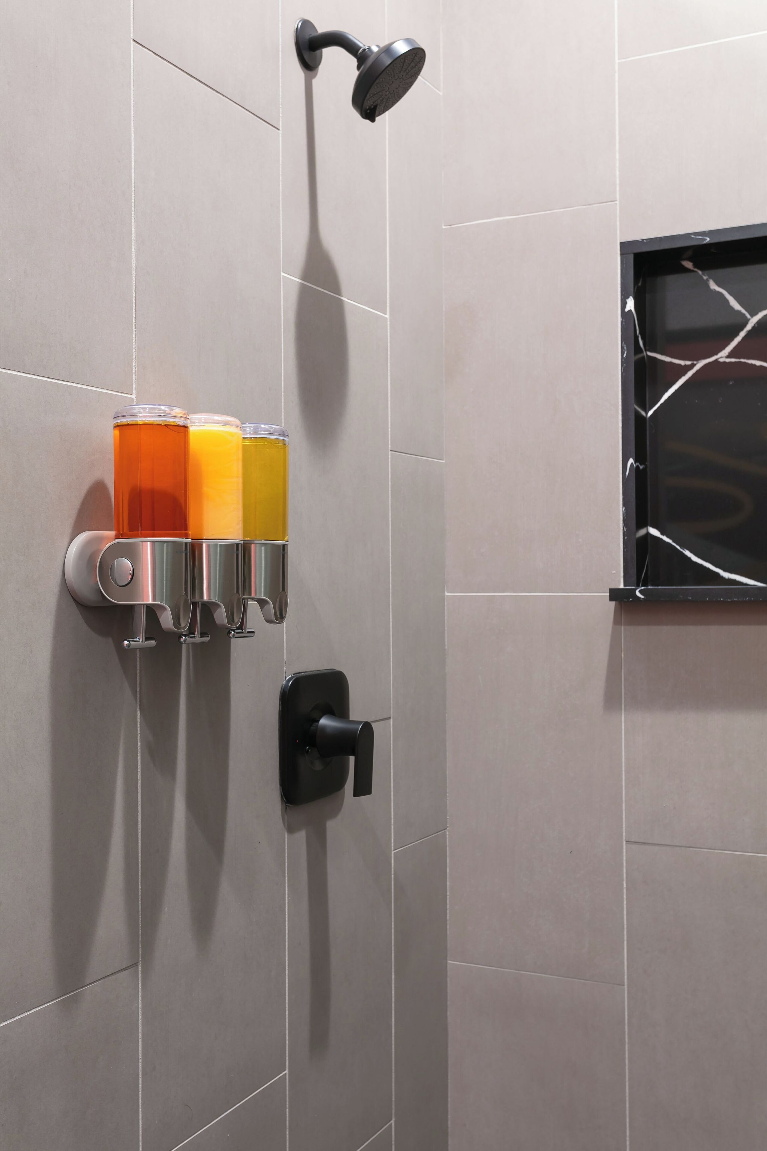 Une salle de bain avec une douche, un porte-bouteilles en verre contenant trois bouteilles et une fenêtre dont le cadre est cassé.