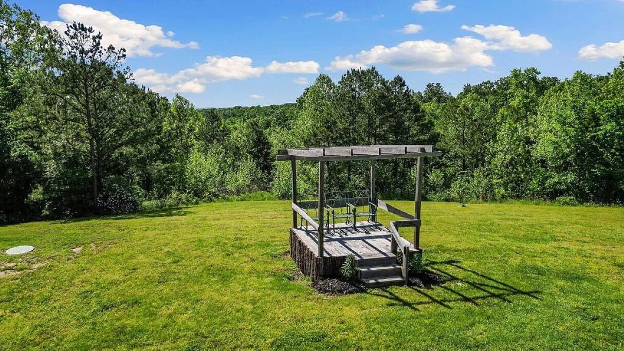 A wooden gazebo in a grassy field with a forest in the background.