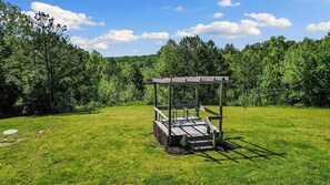 A wooden gazebo in a grassy field with a forest in the background.