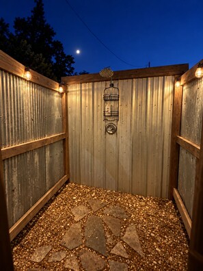 A wooden outdoor shower area with a corrugated metal wall, a stone floor, and a metal rack with bottles.