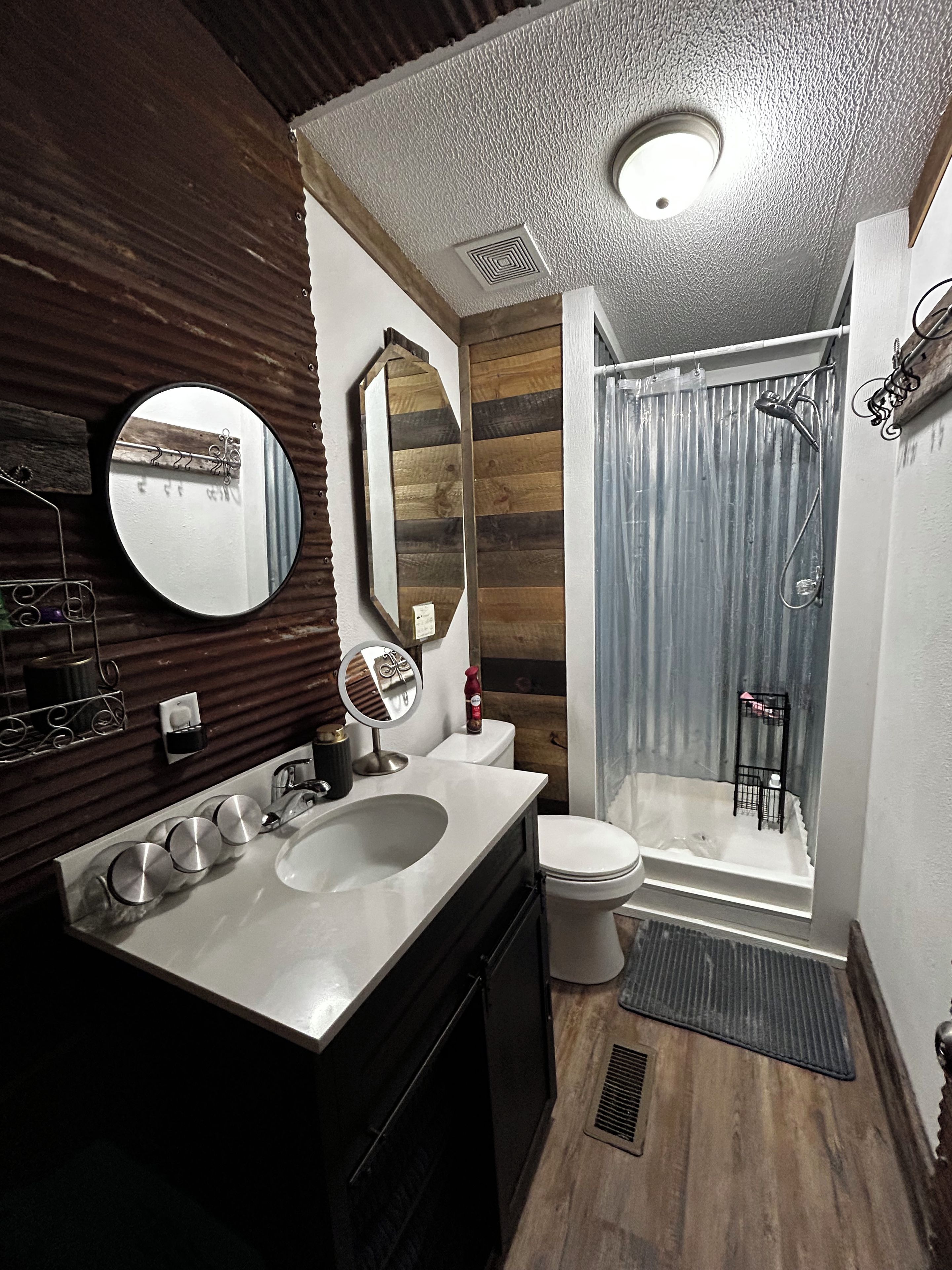 A bathroom with a wooden vanity, a round mirror, a toilet, and a shower with a curtain.