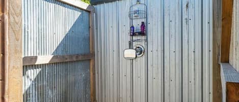 A metal-walled outdoor shower area with a hose, a metal rack with bottles, and a potted plant.