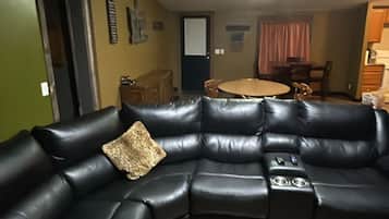 En stue med en sort lædersofa, trægulv, et spisebord med stole og et køkken i baggrunden.