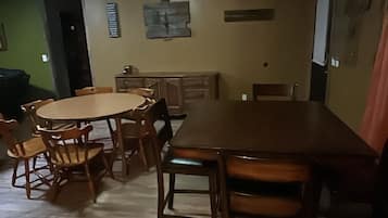 A dining area with a wooden table and chairs, a kitchen in the background, and a green wall.