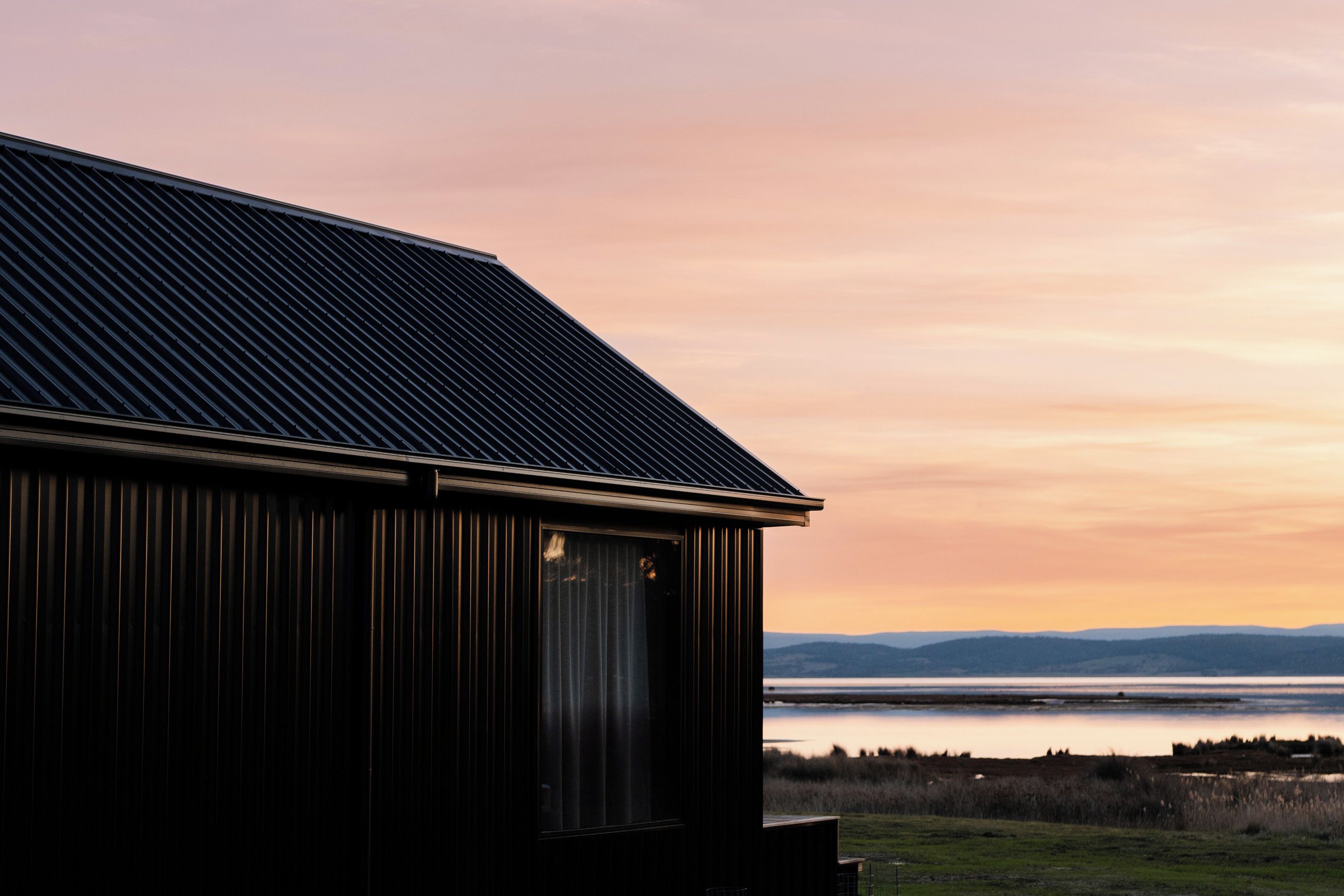 A building with a sloped roof and a large window, overlooking a body of water and distant hills.