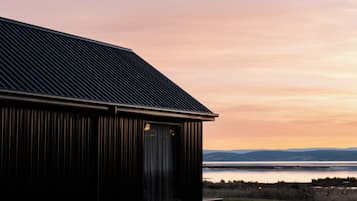 A building with a sloped roof and a large window, overlooking a body of water and distant hills.