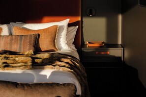 Een net opgemaakt bed met een gestreept deken en kussens, een nachtkastje met een lederen etui, en een wandlamp.