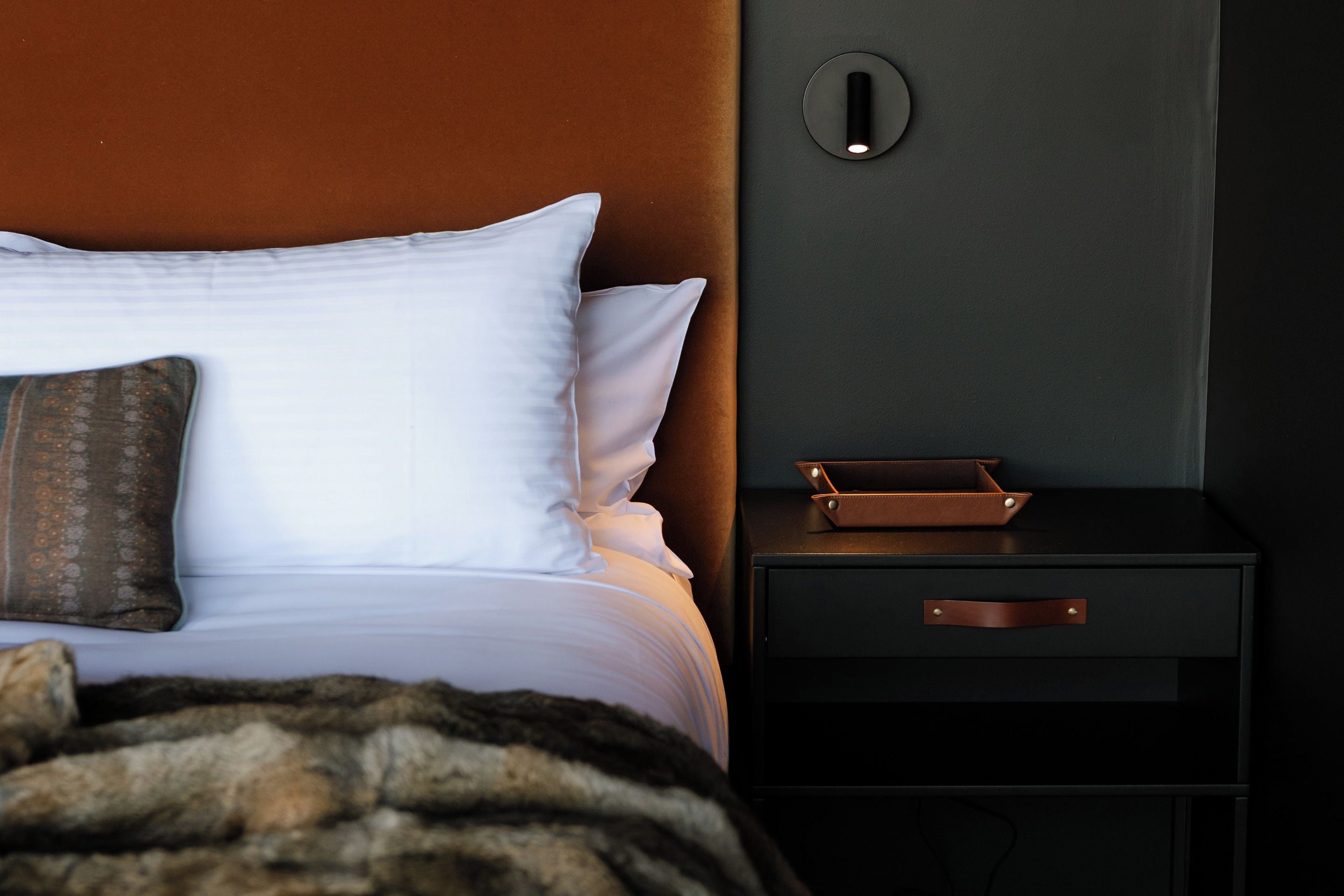 A neatly made bed with white bedding and a brown pillow, a black nightstand with a leather wallet, and a wall-mounted light fixture.