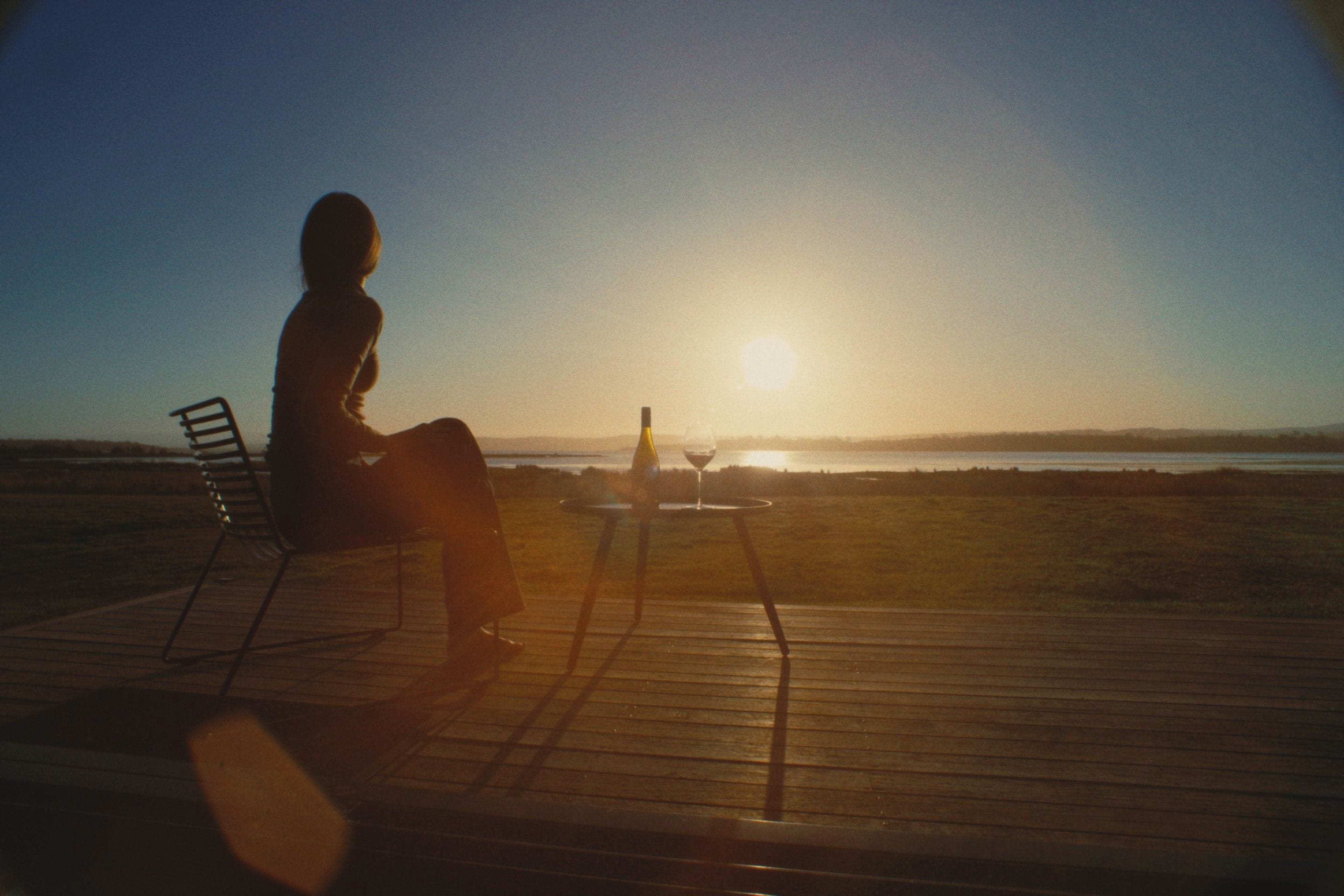 A person sitting on a chair on a wooden deck overlooking a body of water during sunset.