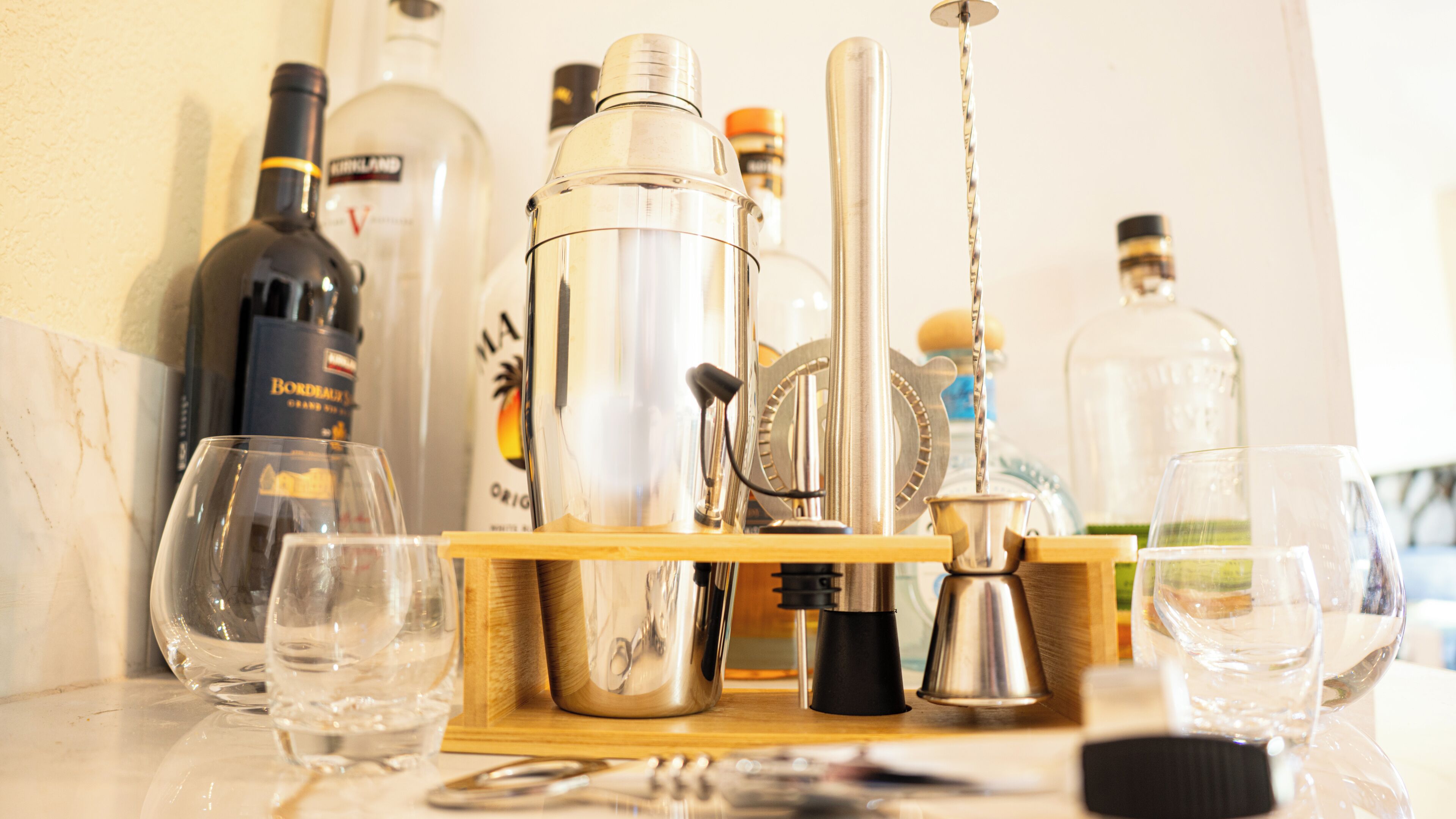 A bar setup with various bottles, a cocktail shaker, and glasses on a wooden tray.