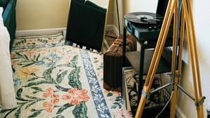 Huoneessa on suuri peili, joka on nojallaan seinää vasten, lattiavalaisin, matkalaukku ja pienellä pöydällä oleva levysoitin.