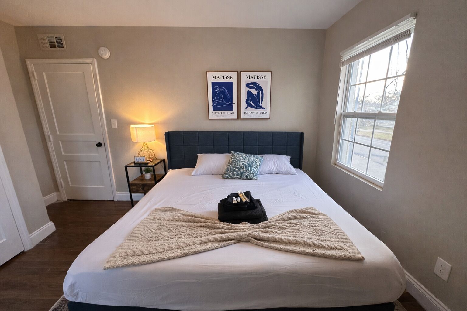 A bedroom with a bed, two wall-mounted artworks, a bedside lamp, and a window.