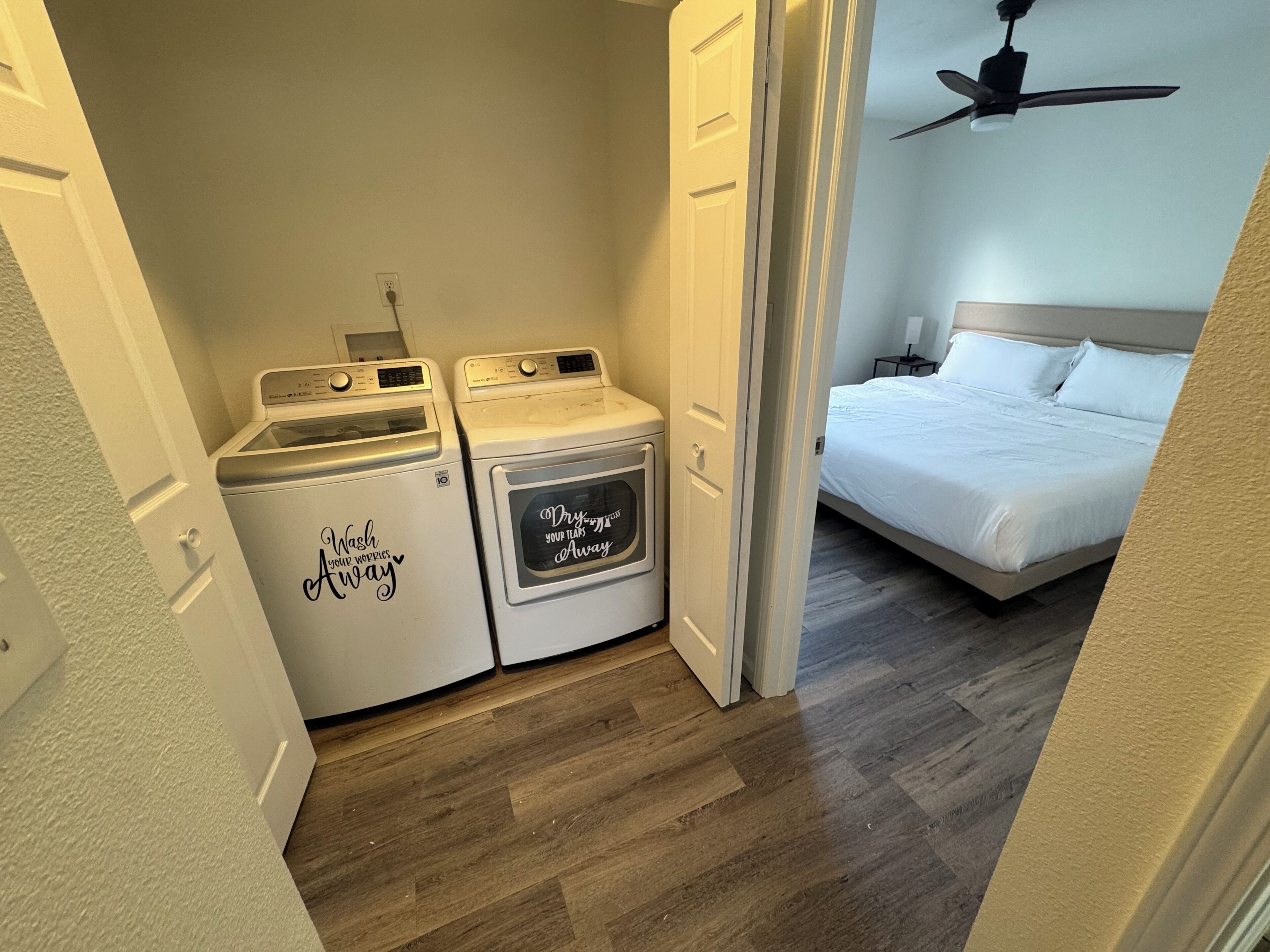A laundry area with two washing machines and a dryer, a bedroom with a bed and a ceiling fan, and wooden flooring.