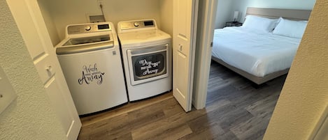 A laundry area with two washing machines and a dryer, a bedroom with a bed and a ceiling fan, and wooden flooring.