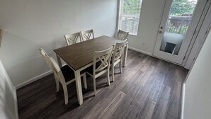 Een eetruimte met een tafel en stoelen, een raam met zonwering, en een deur naar buiten.