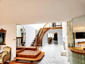 Amplia sala de estar con una escalera de madera que conduce a un nivel superior, un espejo grande y un televisor sobre un mueble verde.