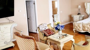 A living room with a wooden floor, a television, a white dresser, a patterned armchair, a glass coffee table, and a bed with a white headboard. - Beautiful 5-bedroom house with AC in wonderful Bay Shore (Bay Shore)
