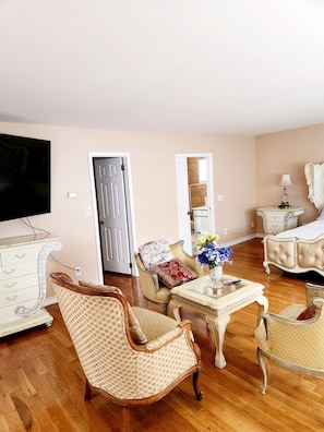 A living room with a wooden floor, a television, a white dresser, a patterned armchair, a glass coffee table, and a bed with a white headboard.