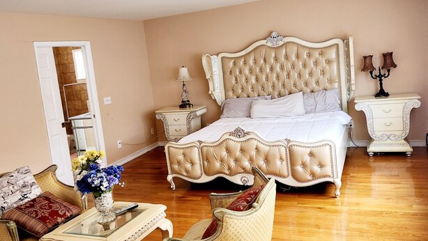 A bedroom with a large bed, a nightstand, a dresser, a chair, and a small table with a vase of flowers. - Beautiful 5-bedroom house with AC in wonderful Bay Shore (Bay Shore)