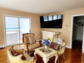A living room with a television mounted on the wall, a glass coffee table with a vase of flowers, and two armchairs with cushions.
