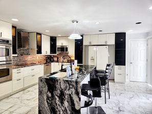 A modern kitchen with white cabinets, a granite countertop, a stainless steel oven, a microwave, a refrigerator, and a bar area with stools.