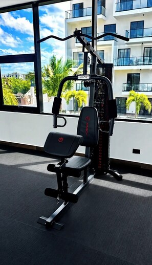 Ein Fitnessraum mit Langhantelbank und Gewichtsblock, große Fenster mit Blick auf ein Gebäude und Palmen.