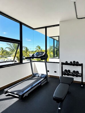 Une salle de sport équipée d’un tapis de course, d’un banc de musculation et d’haltères, avec de grandes fenêtres donnant sur des palmiers.