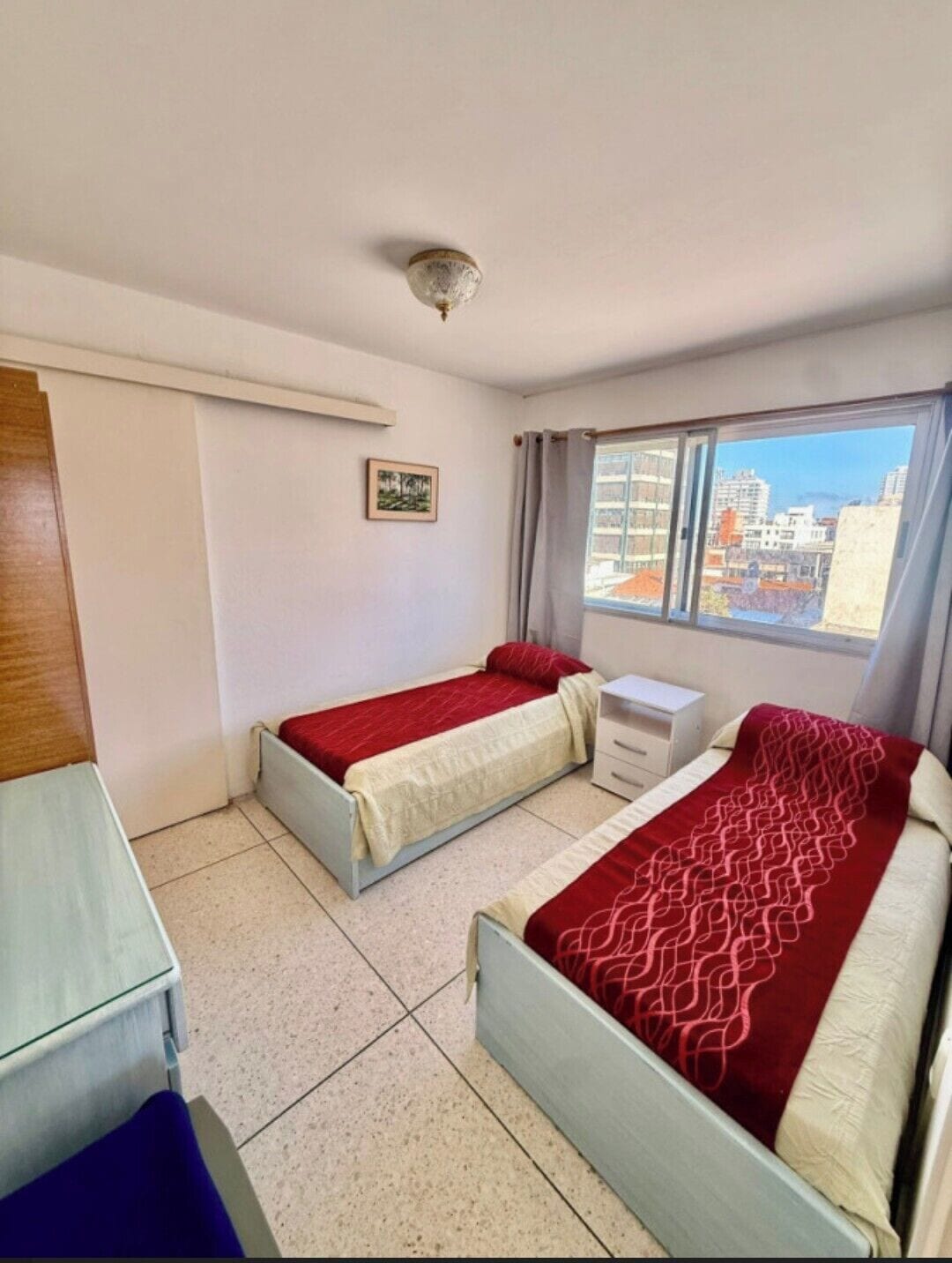 A room with two beds, a small desk, a window with curtains, and a ceiling light.