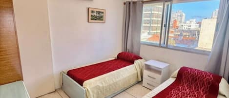 A room with two beds, a small desk, a window with curtains, and a ceiling light.