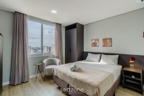 A hotel room with a large bed, a chair, a nightstand, a window with curtains, and a city view. - Studio in Historic Center Near Market Tco1106 (Porto Alegre)