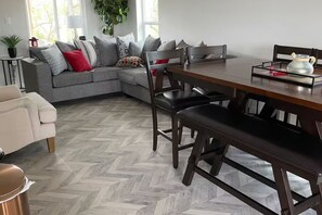 A modern living room with a herringbone floor, a grey sectional sofa with patterned cushions, a wooden dining table with chairs, and a small side table with a lamp and decorative items. - Entire home: Residential home in Rio Hondo, Texas, United States. (Rio Hondo)