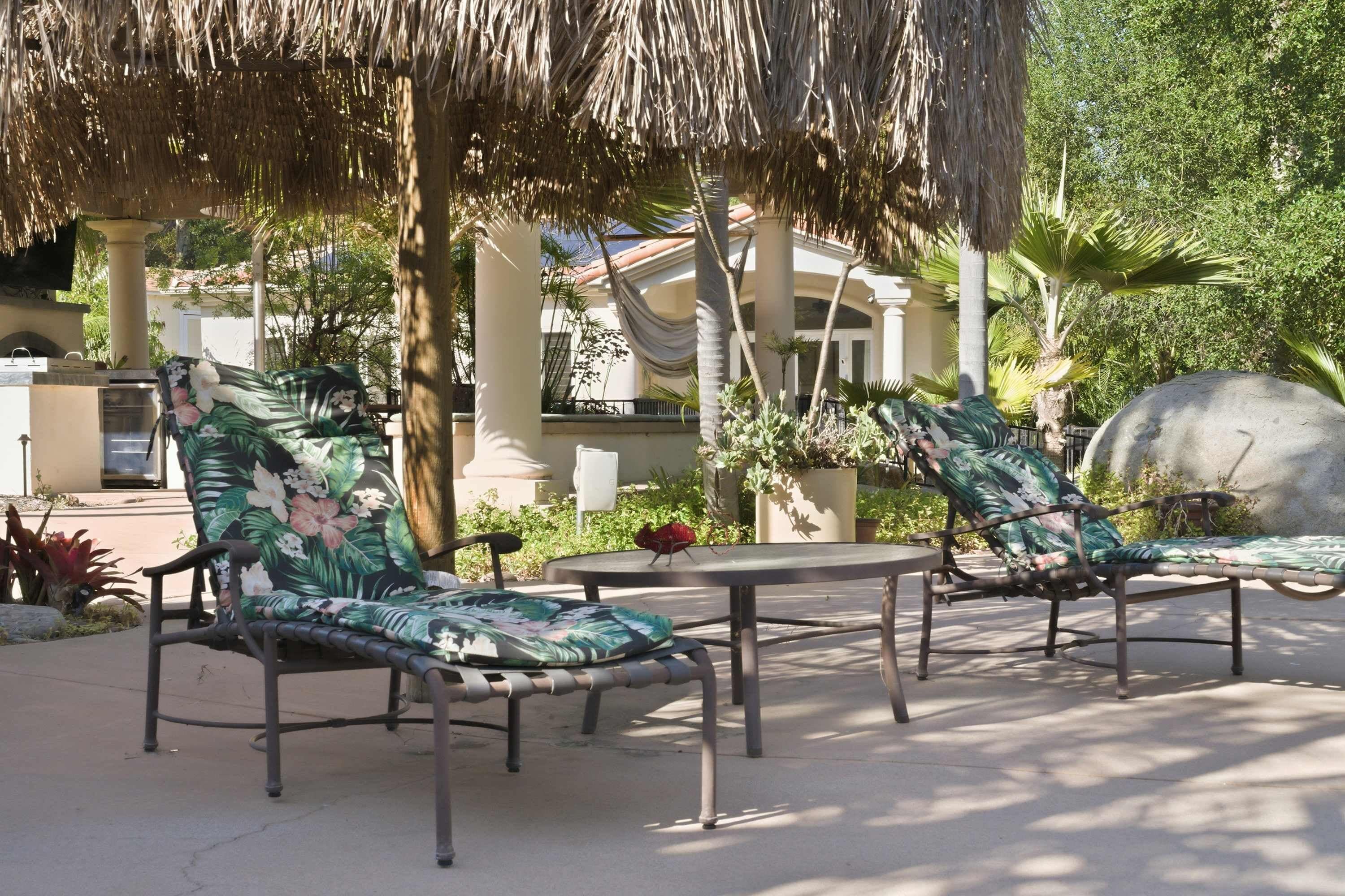 An outdoor seating area with two lounge chairs and a table, surrounded by tropical plants and a thatched roof structure.