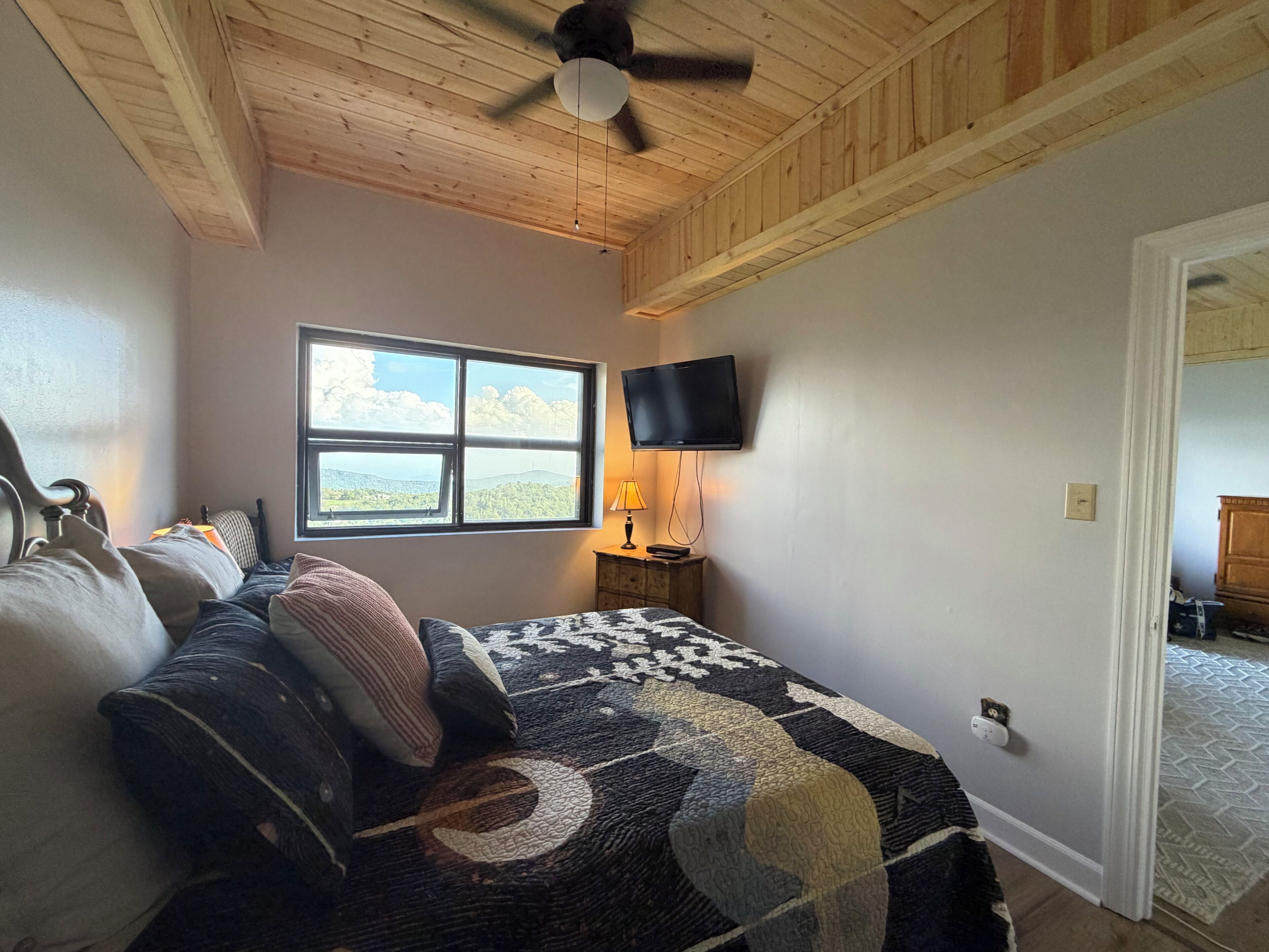 A bedroom with a bed, a ceiling fan, a TV mounted on the wall, and a window with a view of the outdoors.